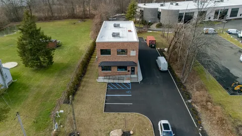 Aerial view of the brick office with its flat white roof, paved drive and parking, a lawn, and a larger commercial building beside it